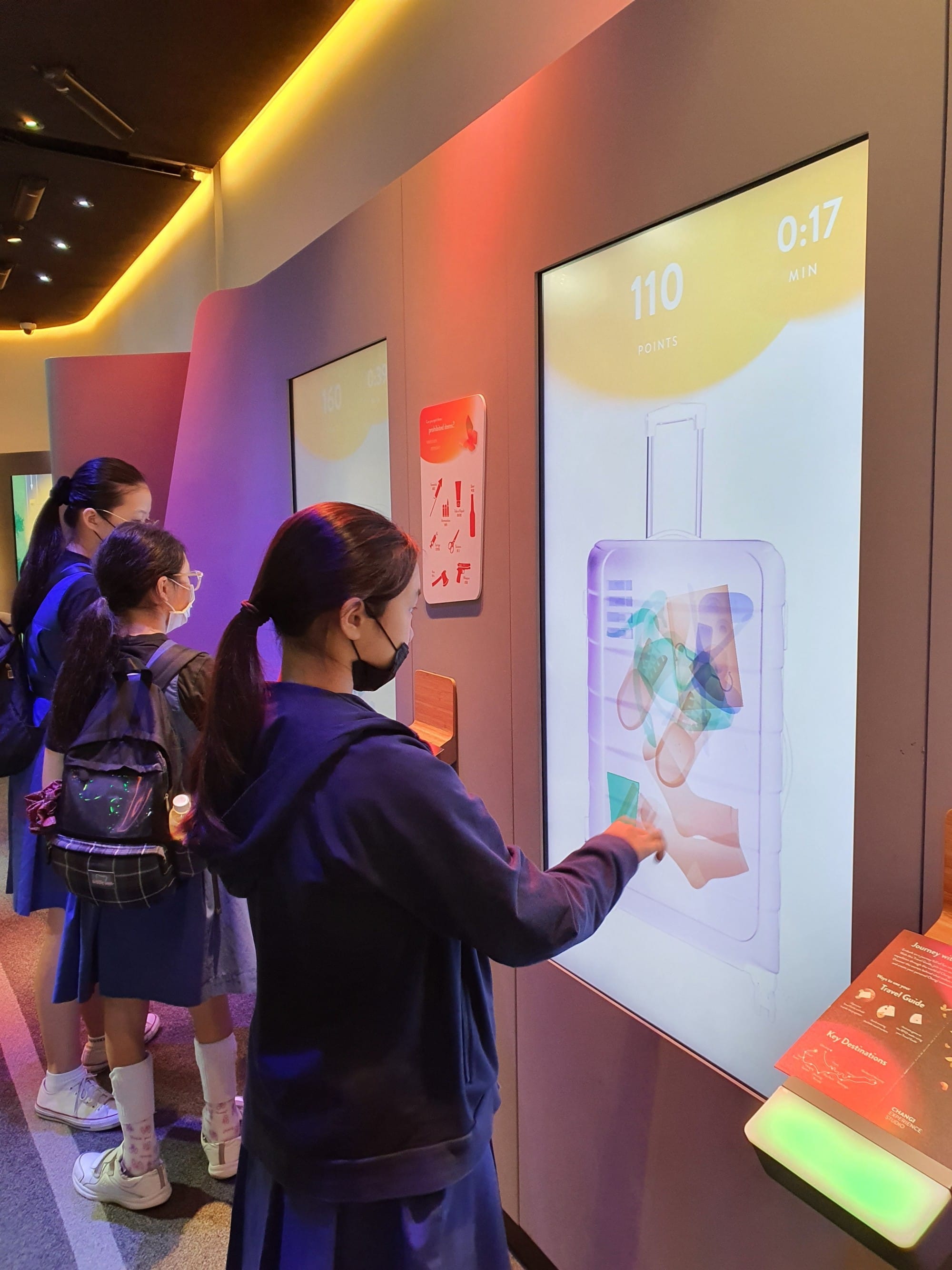 Three masked girls interact with a large touchscreen showing an X-ray luggage scanning game with 110 points displayed.